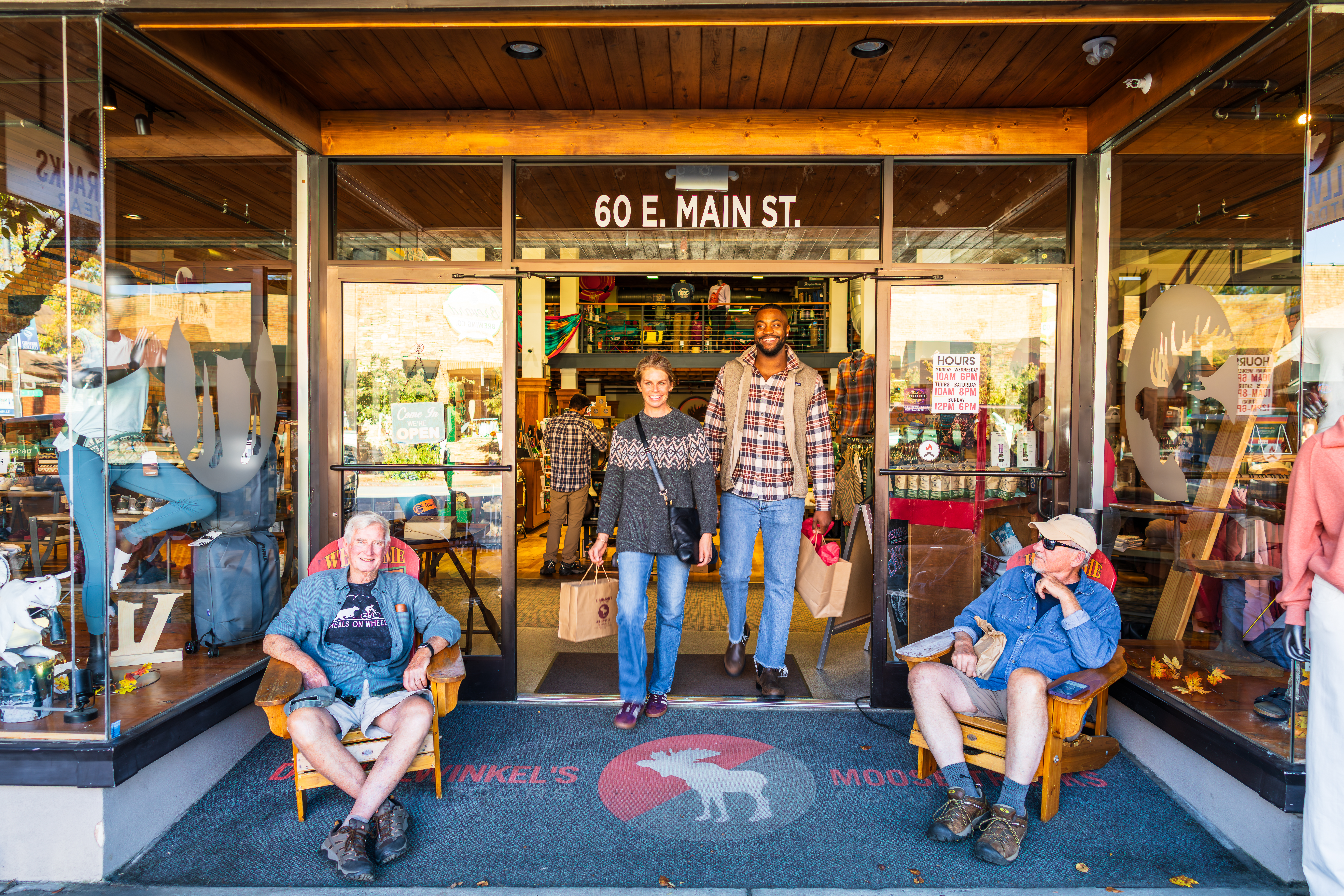 A man and woman walking out of D.D. Bullwinkel's Outdoors with shopping bags. Two people resting on chairs outside the door in Downtown Brevard, NC