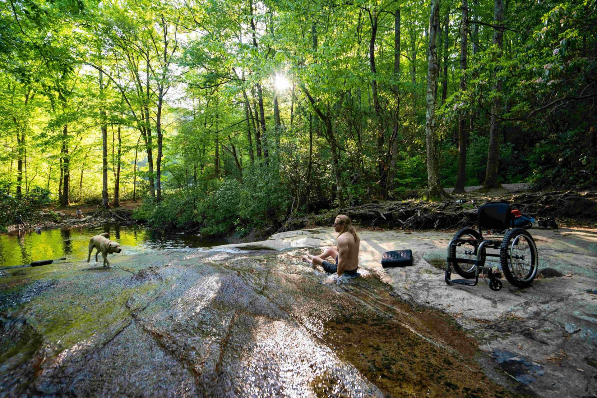 Man sitting in the water next to a wheelchair, watching his dog play in the river.